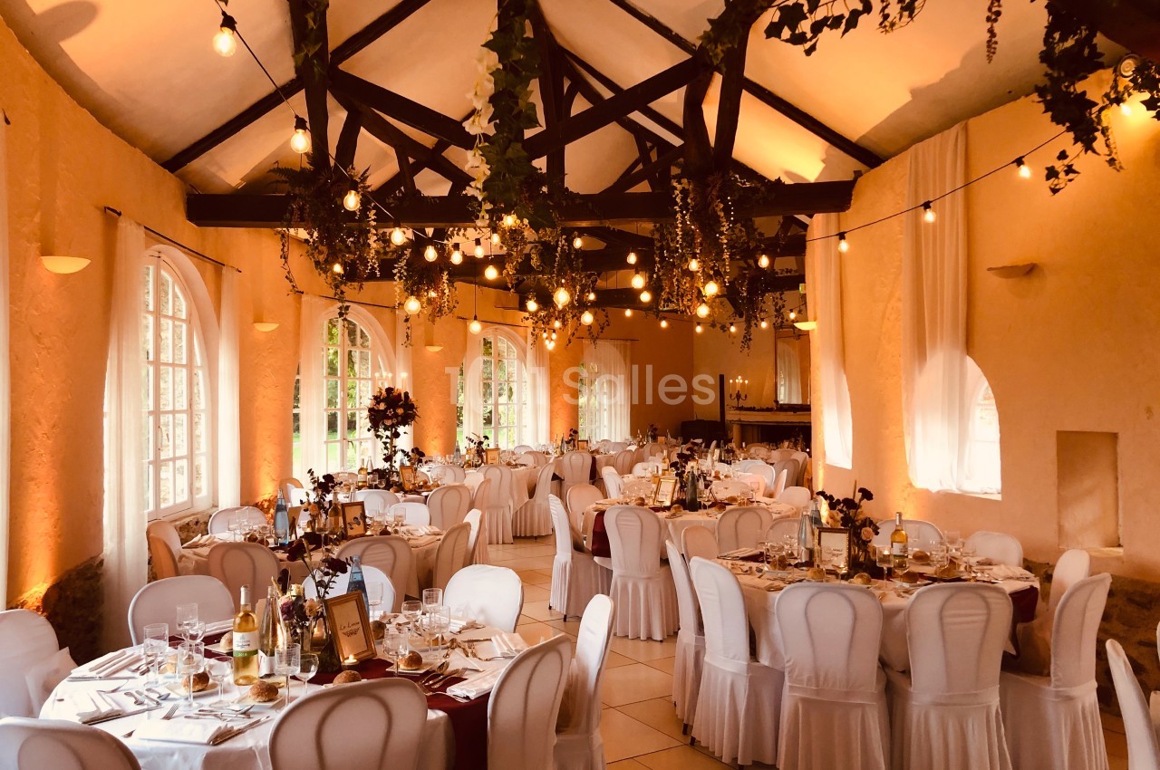Salle de réception décorée avec des guirlandes lumineuses, des tables dressées et des chaises blanches.