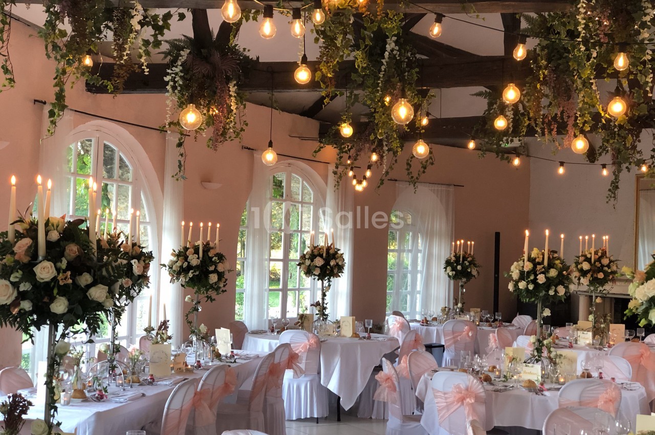 Salle de réception décorée pour un mariage avec guirlandes lumineuses, fleurs suspendues et tables élégamment dressées.