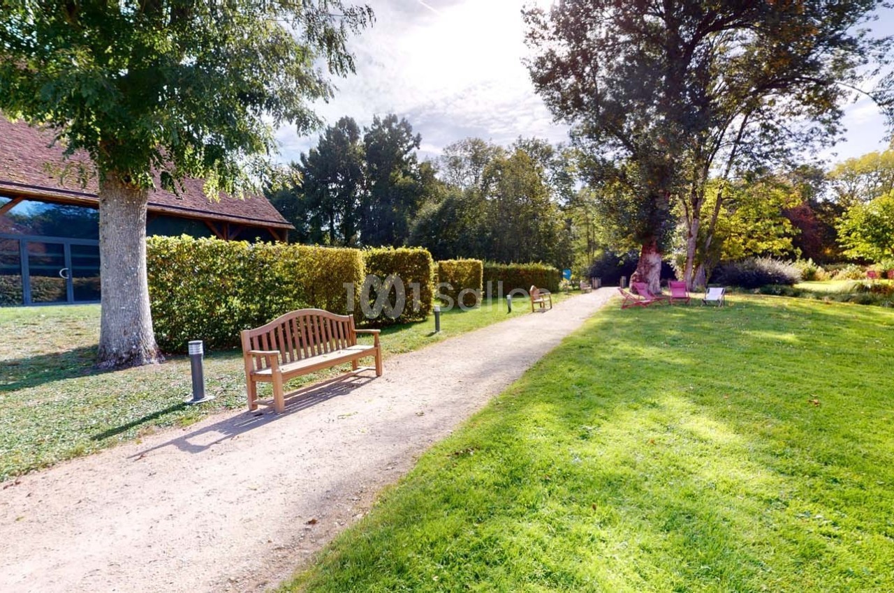 Allée bordée de bancs en bois, haies taillées et pelouse, menant à un bâtiment avec toit en pente sous un ciel dégagé.