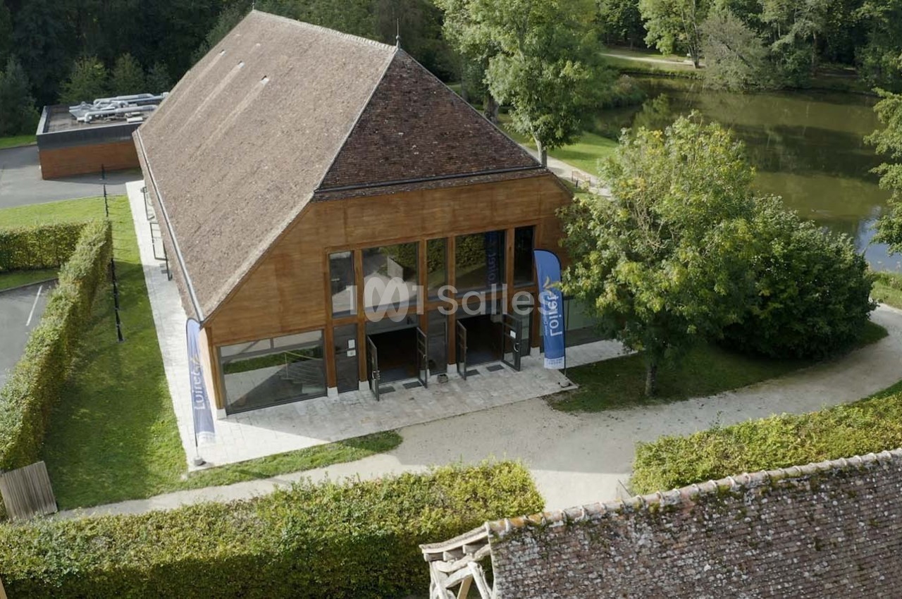 Bâtiment en bois avec de grandes baies vitrées, entouré de verdure, près d'un étang et d'un parking.