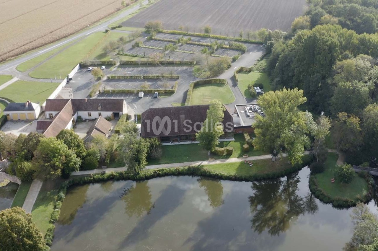 Vue aérienne d'un domaine avec bâtiments, étang, parking et champs environnants, entouré de végétation.