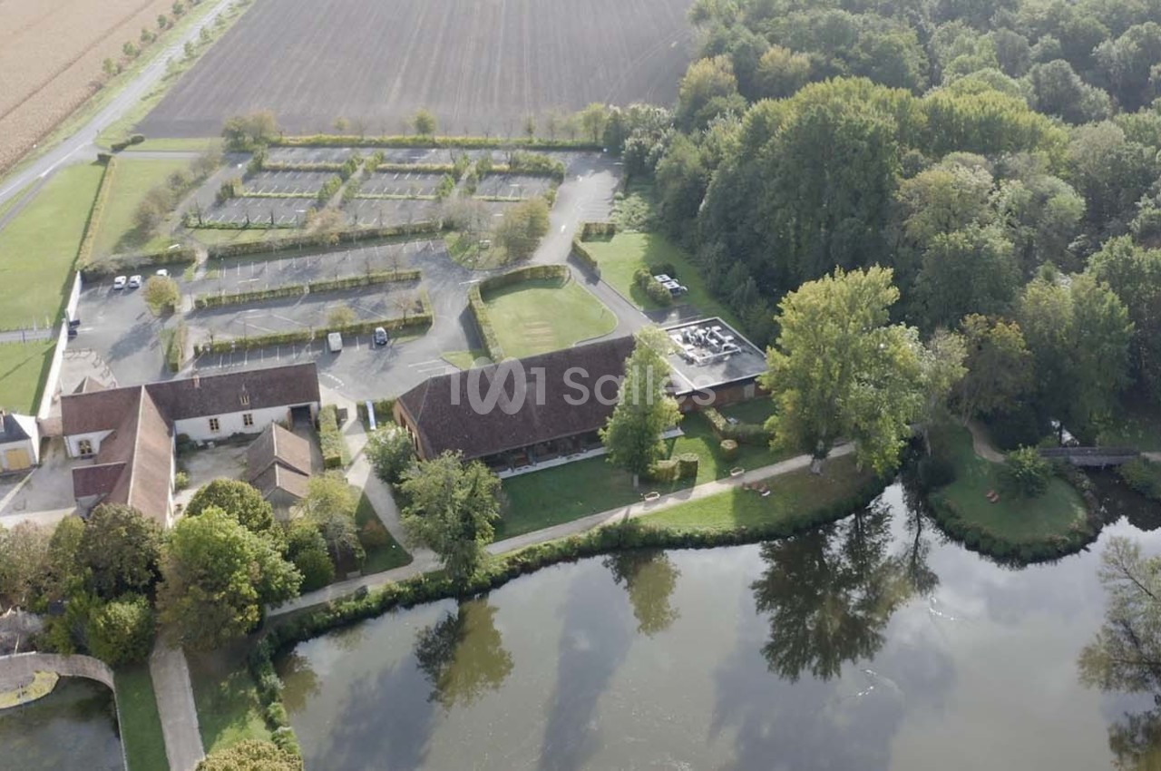 Vue aérienne d'un domaine rural avec bâtiments, parking, étang et forêt environnante.