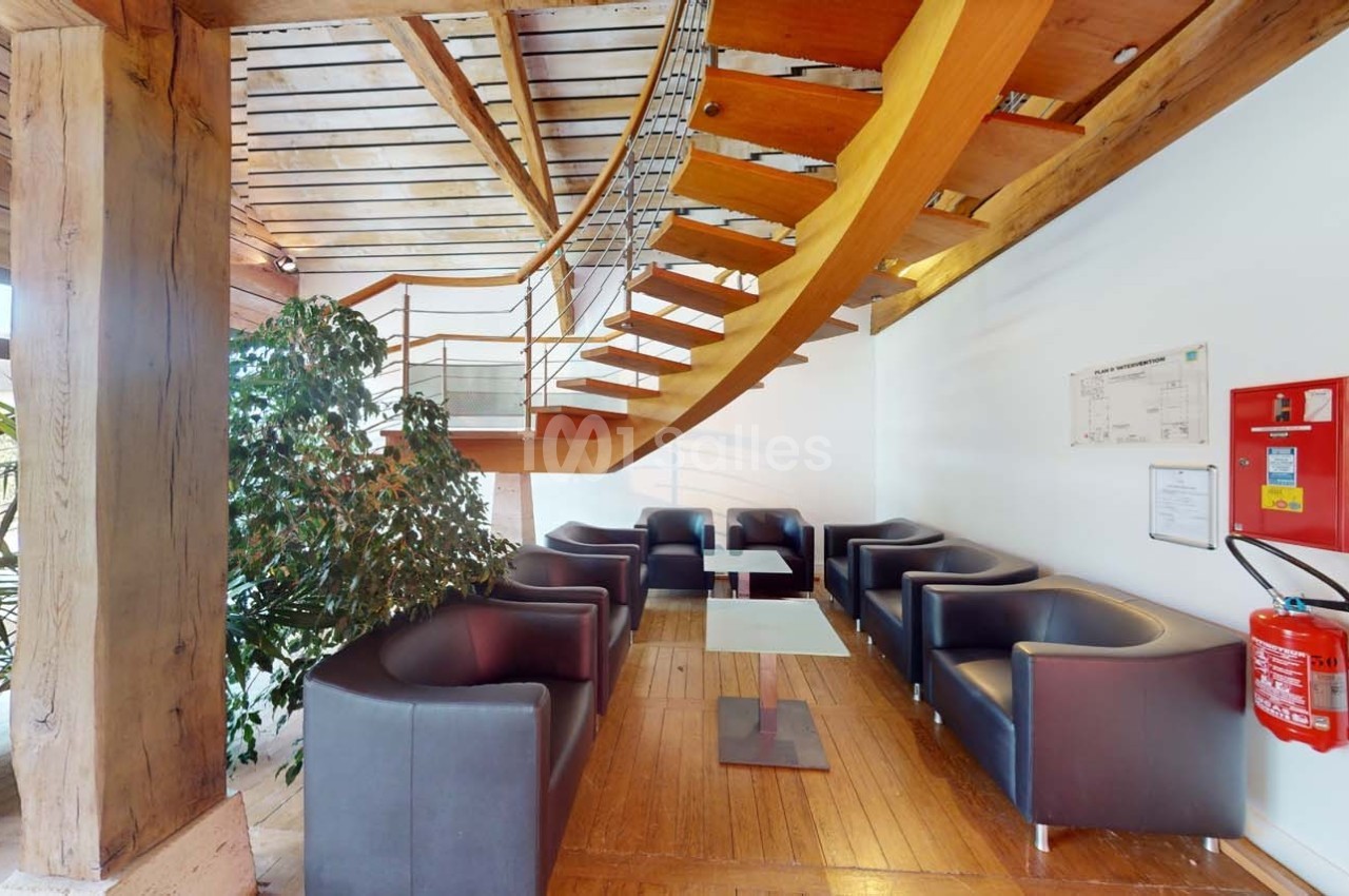 Espace d'attente avec fauteuils noirs, tables basses, escalier en bois courbé et plantes dans un intérieur lumineux.