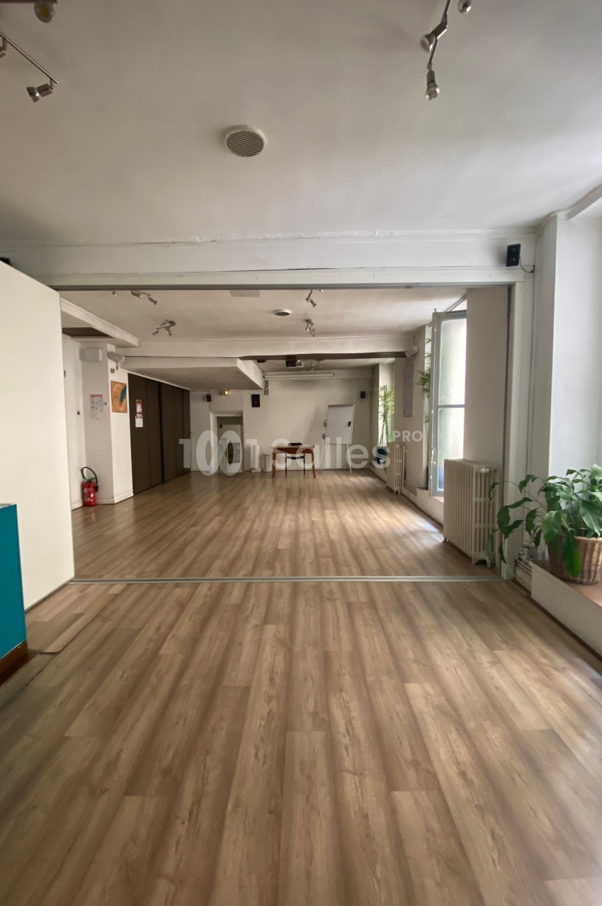 Salle lumineuse avec parquet clair, murs blancs, grandes fenêtres, plantes en pot et mobilier minimaliste.