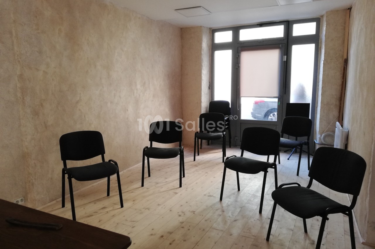 Salle lumineuse avec murs clairs, parquet en bois et plusieurs chaises noires disposées en cercle.