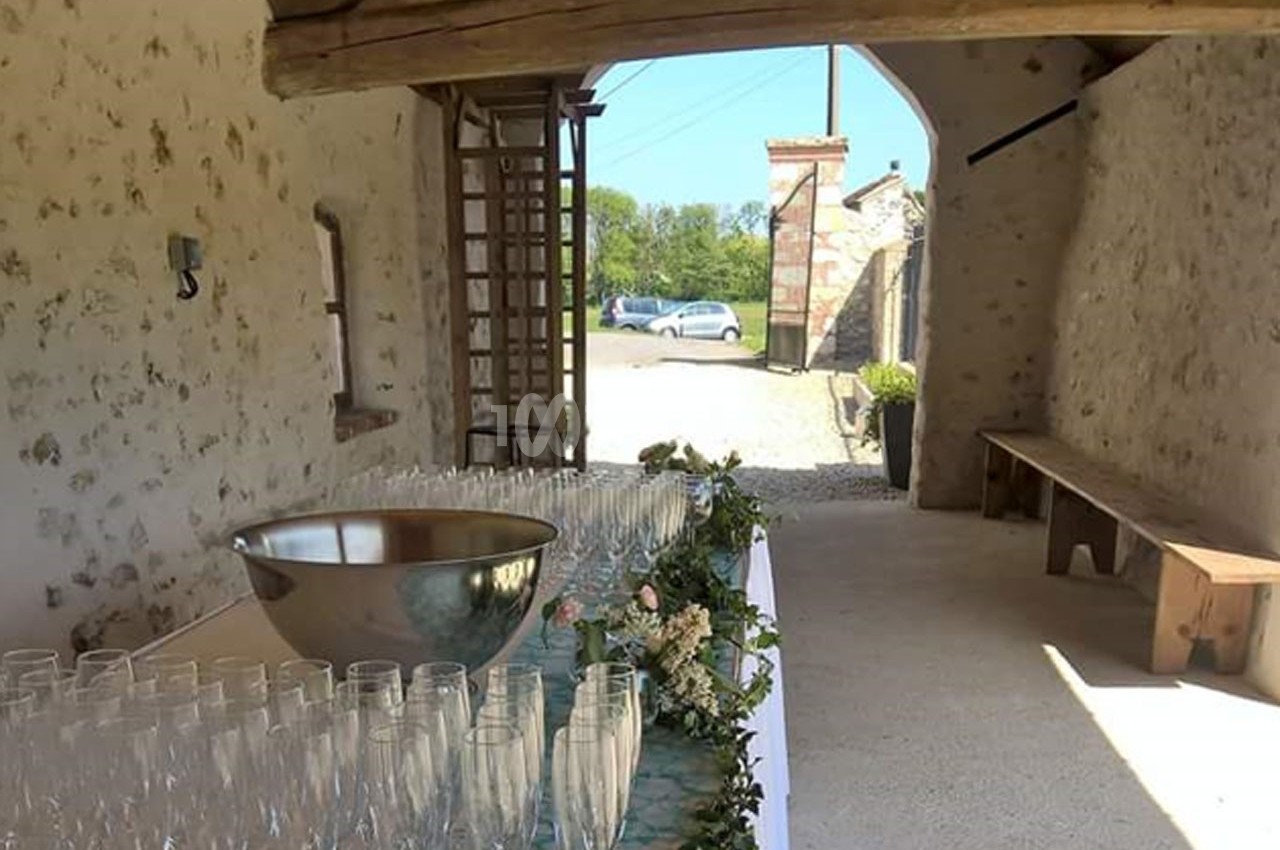 Table décorée avec des verres et une coupe en métal dans une grange ouverte, avec vue sur l'extérieur ensoleillé.