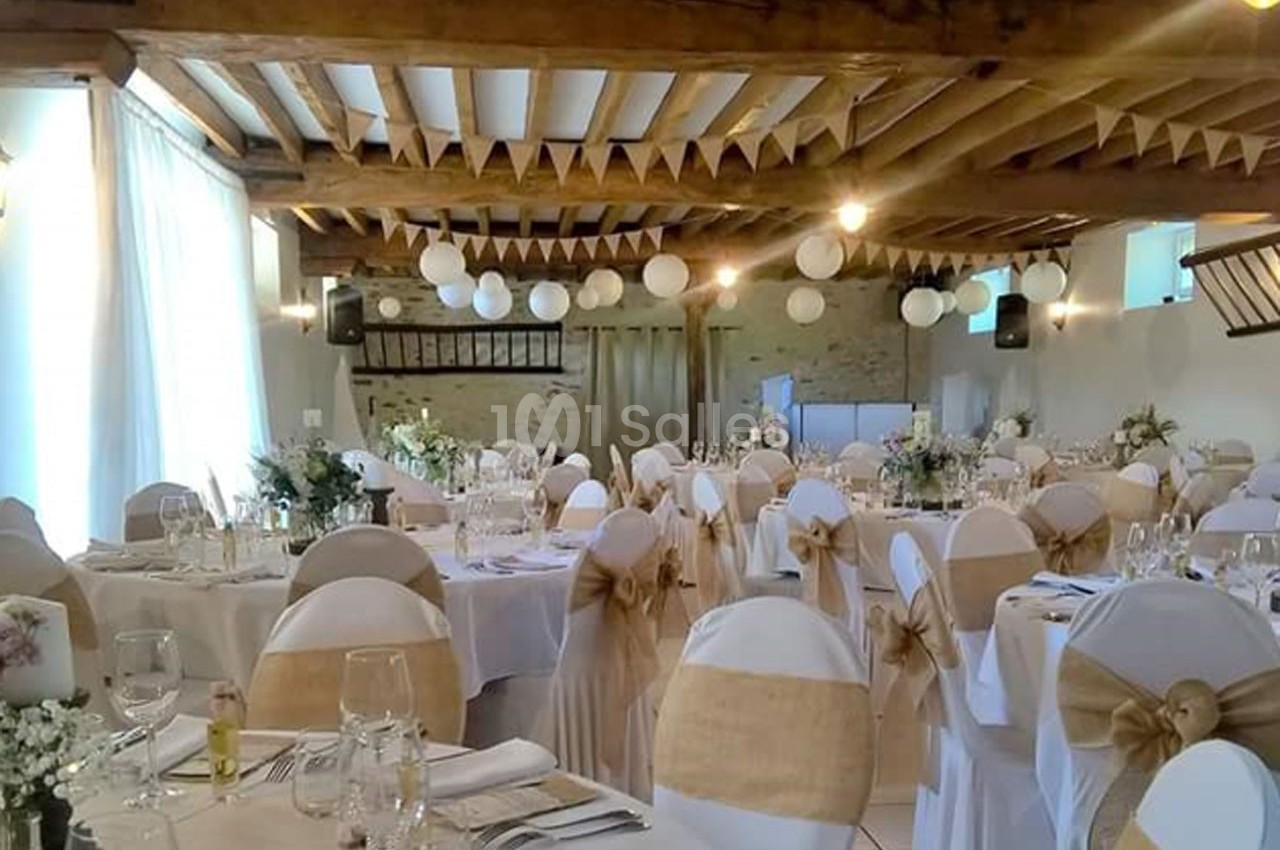 Salle de réception décorée avec des tables rondes, nappes blanches, chaises ornées de nœuds en toile de jute et guirlandes…