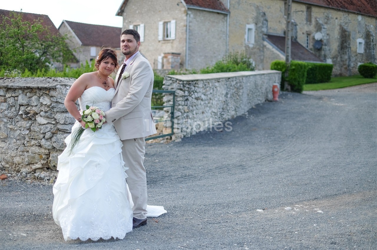 Un couple en tenue de mariage pose devant un mur de pierre, avec des bâtiments en arrière-plan.