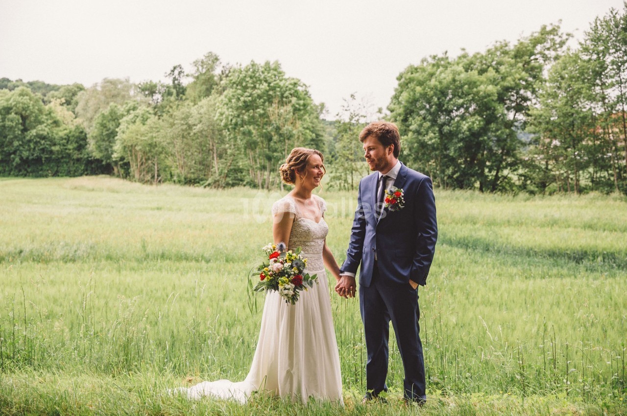 Un couple en tenue de mariage se tient la main dans un champ verdoyant bordé d'arbres.