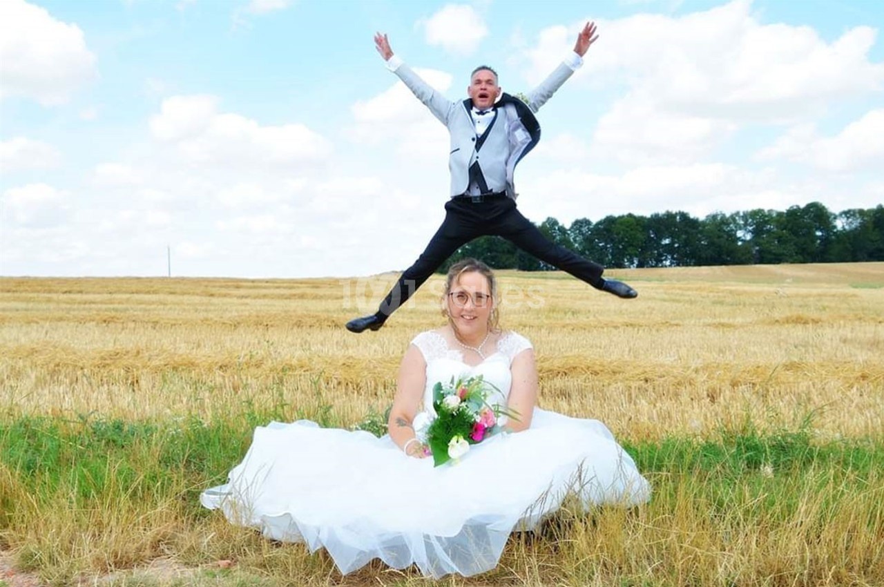 Une mariée assise dans un champ tient un bouquet, tandis qu'une personne en costume saute en arrière-plan.
