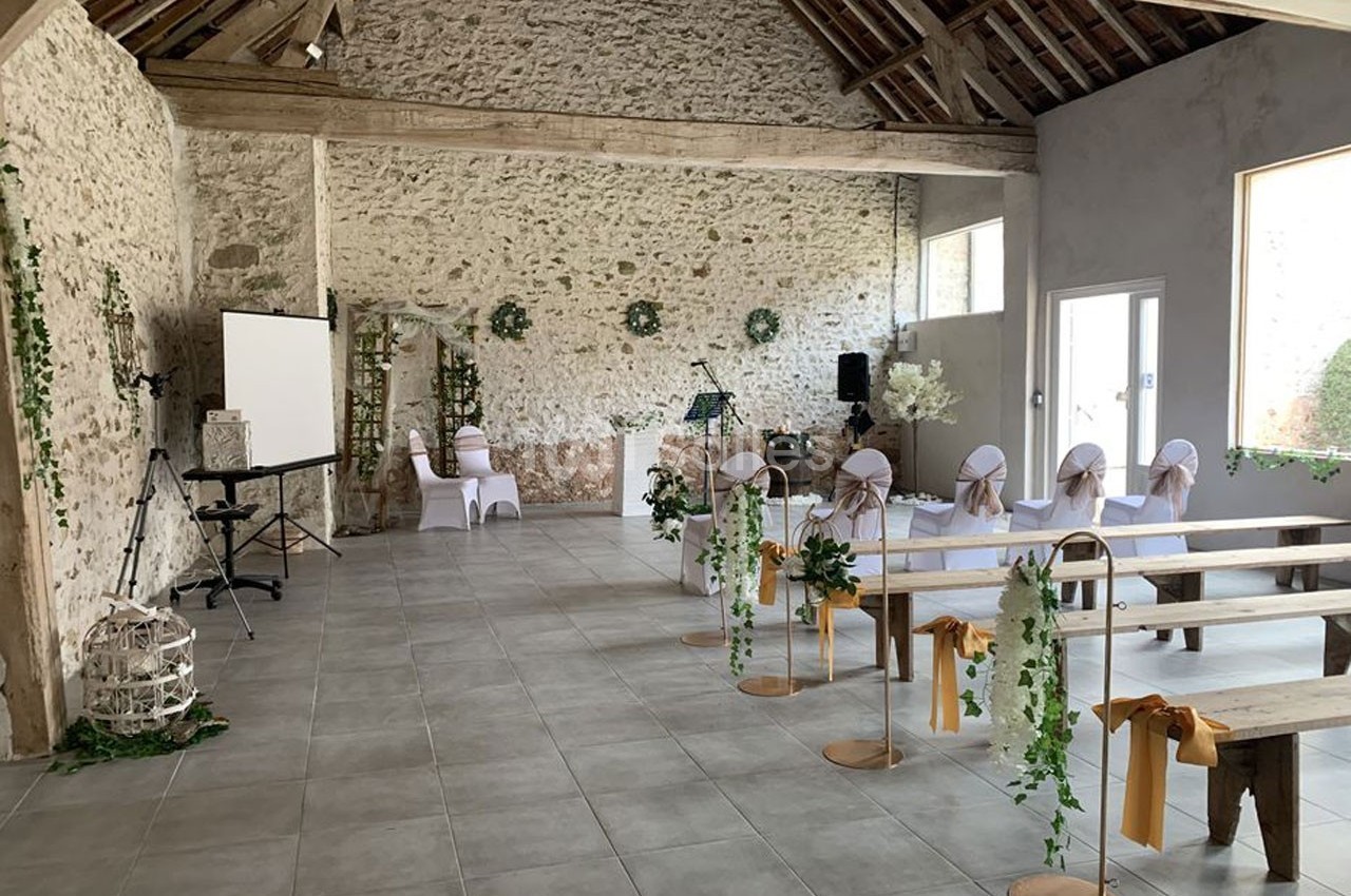 Salle de réception rustique décorée pour un événement avec chaises blanches, fleurs et pupitre, dans un cadre en pierre.