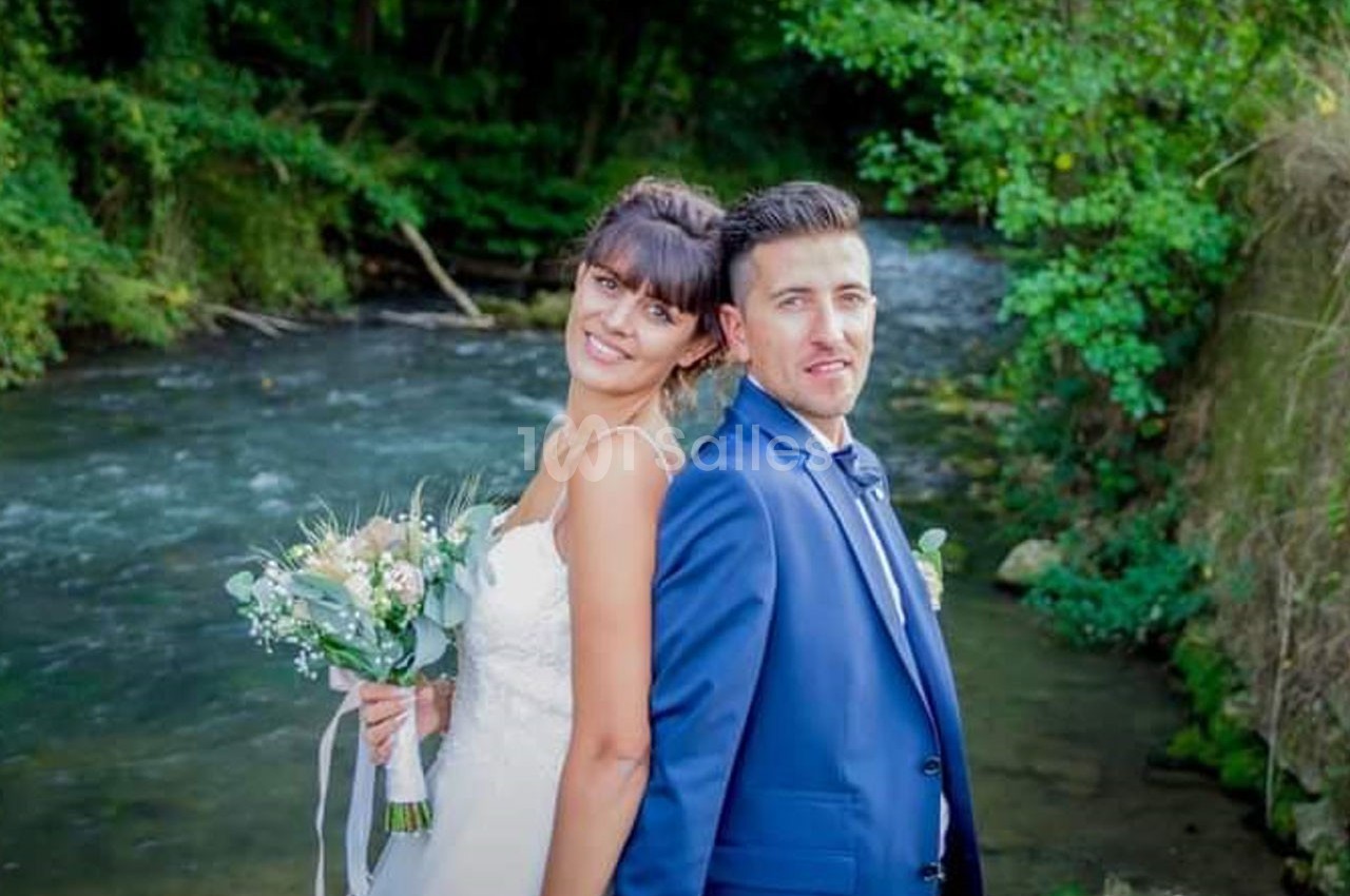 Un couple en tenue de mariage pose dos à dos près d'une rivière entourée de végétation.
