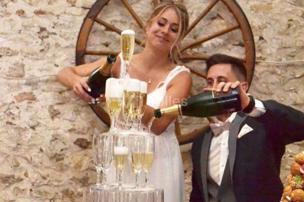 Un couple en tenue de mariage verse du champagne dans une pyramide de coupes devant un mur en pierre.