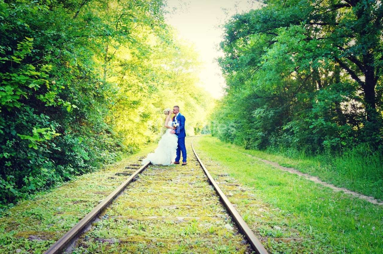 Un couple en tenue de mariage pose sur des rails entourés de verdure dans un cadre naturel et lumineux.