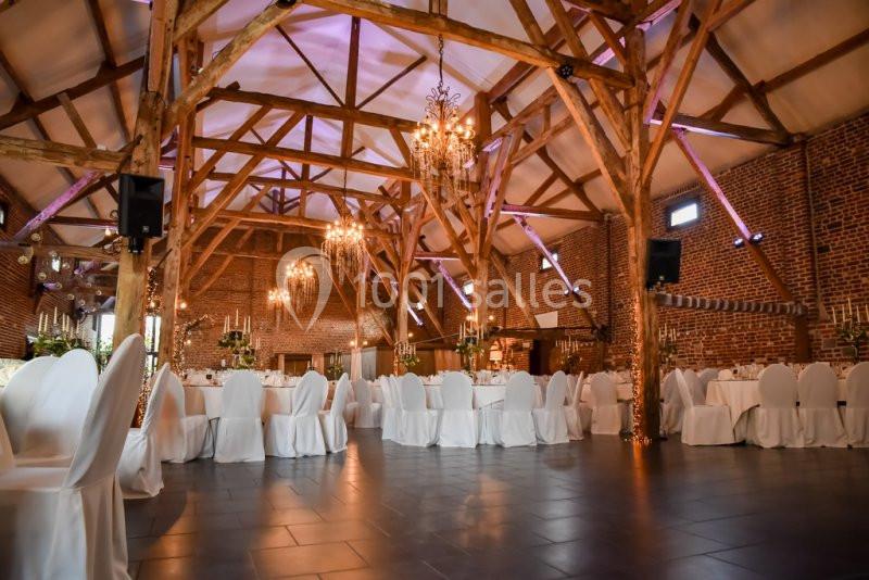 Salle de réception avec poutres apparentes, murs en briques, chaises blanches et lustres suspendus.