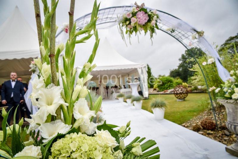 Arrangement floral blanc et vert au premier plan, avec une arche décorée et une tente de réception en arrière-plan.