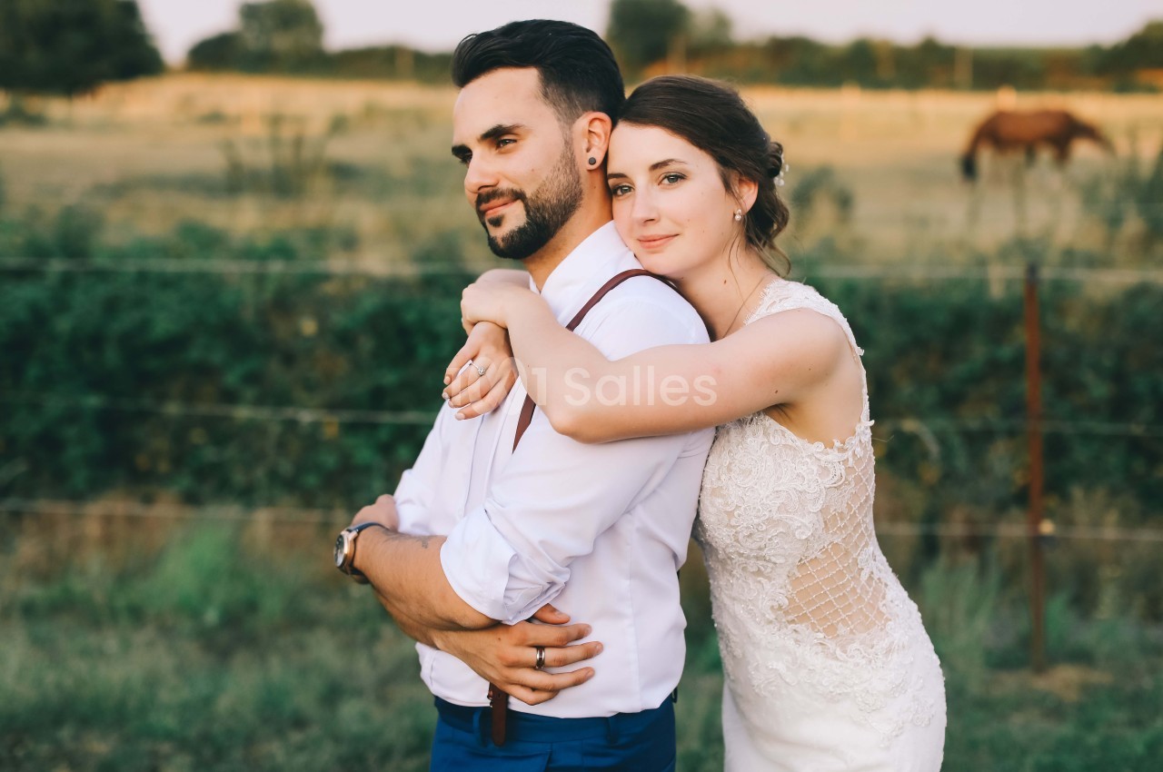 Un couple en tenue de mariage pose dans un champ, la mariée entourant le marié de ses bras.