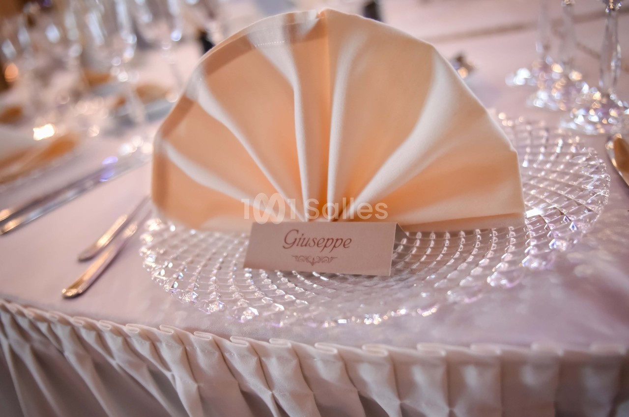 Serviette pliée en éventail sur une assiette en verre, avec un marque-place portant le prénom ’Giuseppe’.