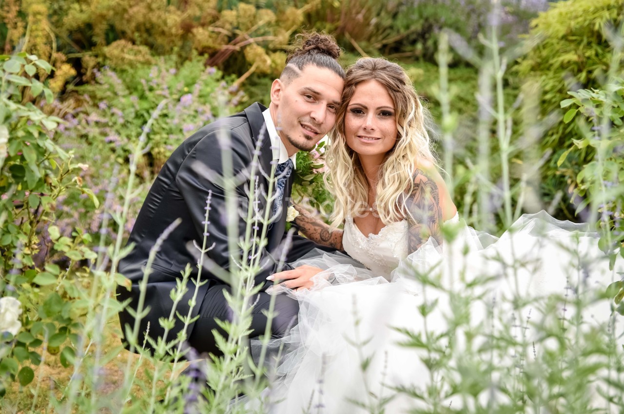 Un couple en tenue de mariage pose dans un jardin verdoyant avec des fleurs en arrière-plan.