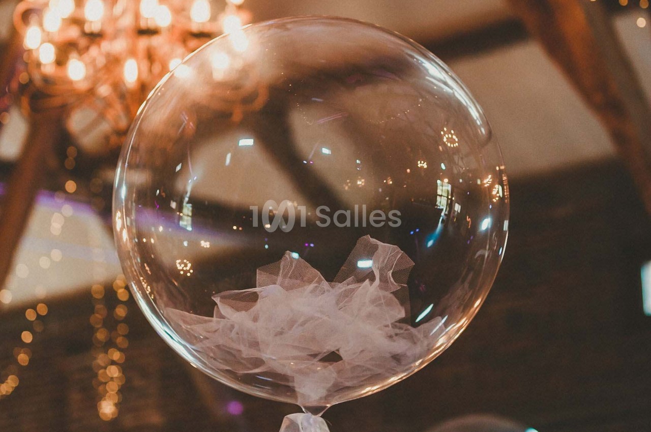 Ballon transparent décoratif avec un nœud en tulle blanc, suspendu sous un lustre dans une salle éclairée.