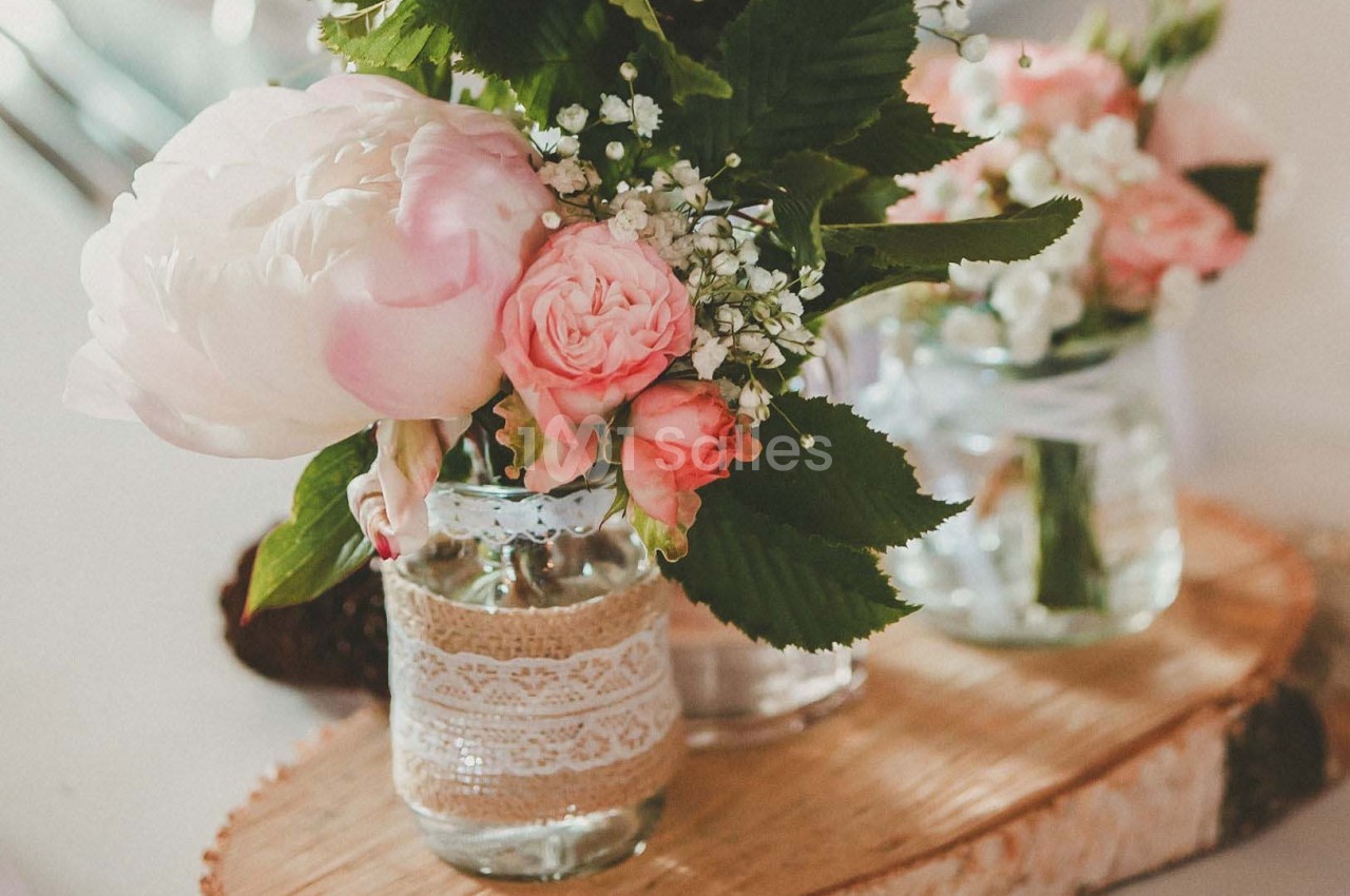 Bouquets de fleurs roses et blanches dans des bocaux en verre décorés de dentelle, posés sur une planche en bois.