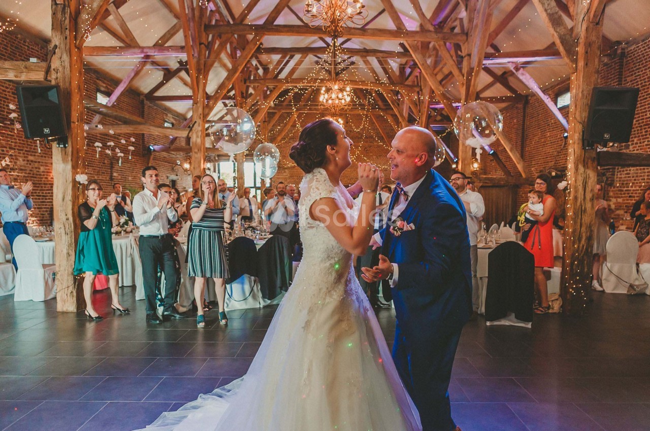 Un couple danse lors d'une réception dans une salle en bois avec des invités applaudissant en arrière-plan.