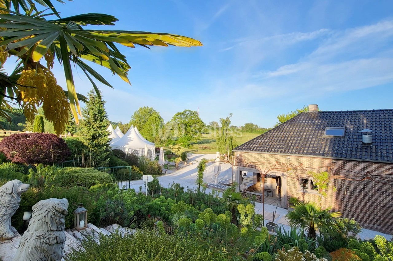 Jardin verdoyant avec une maison en briques, des tentes blanches et un ciel bleu dégagé en arrière-plan.