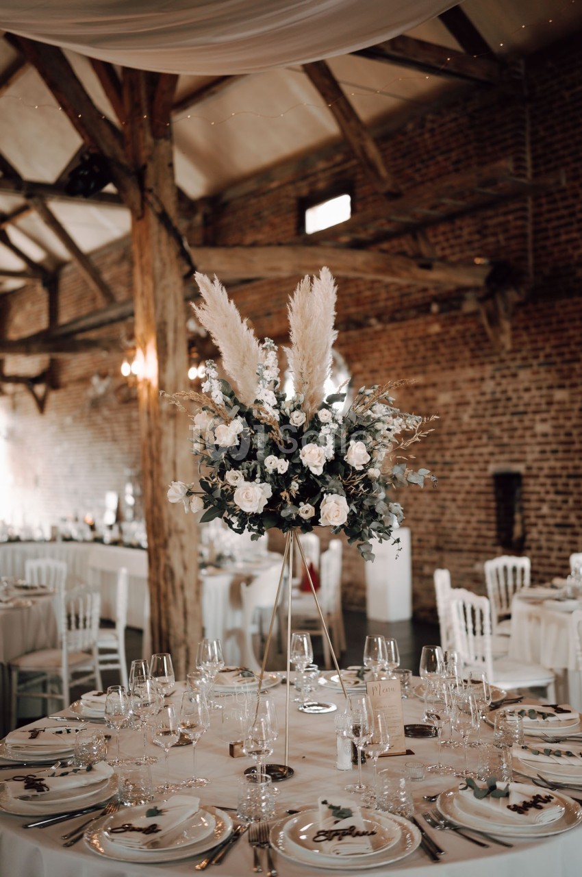 Centre de table floral avec pampas et roses blanches dans une salle de réception en briques avec poutres apparentes.
