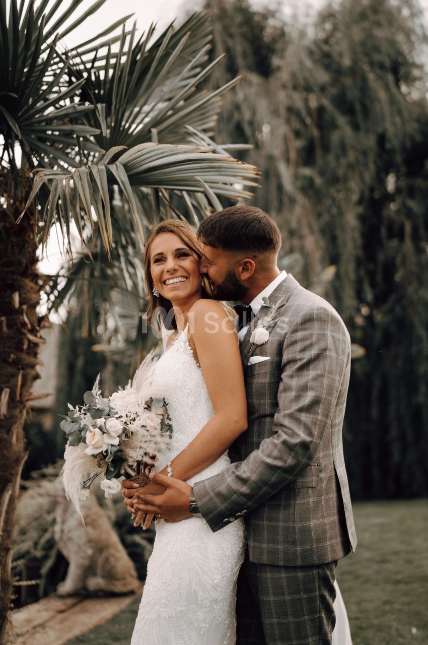 Un couple de mariés souriant, l'homme en costume gris embrasse la femme en robe blanche tenant un bouquet, devant des arbres.