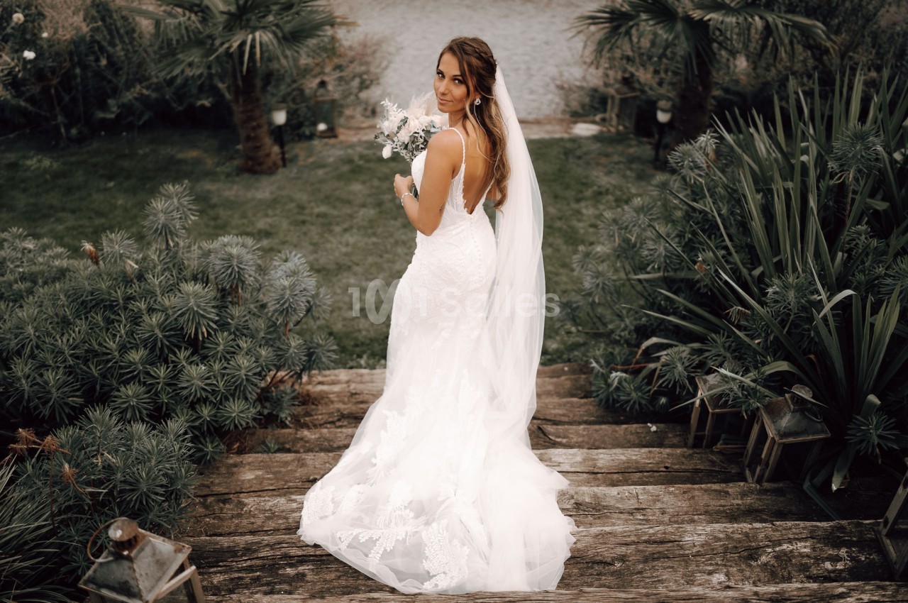 Femme en robe de mariée blanche tenant un bouquet, debout sur des marches en bois entourées de végétation.