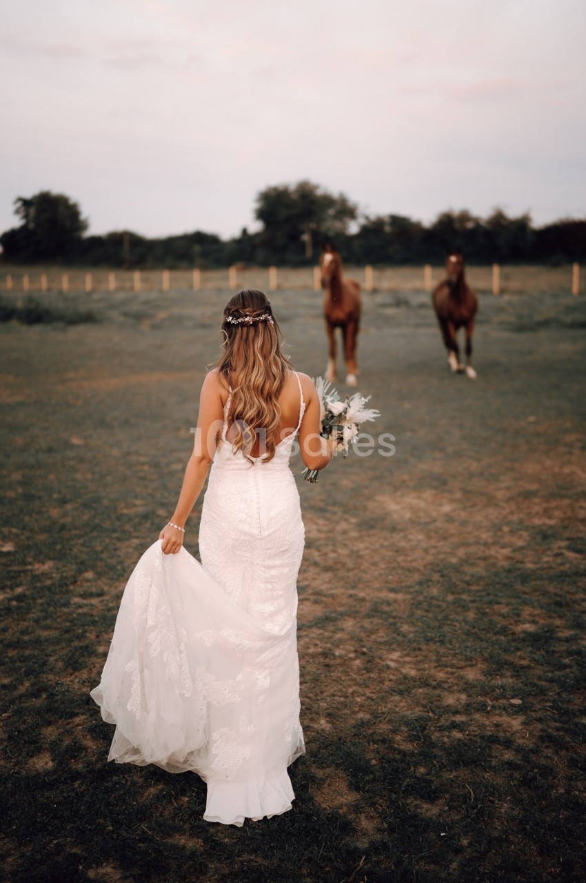 Une femme en robe de mariée marche dans un champ, tenant sa traîne et un bouquet, avec deux chevaux en arrière-plan.