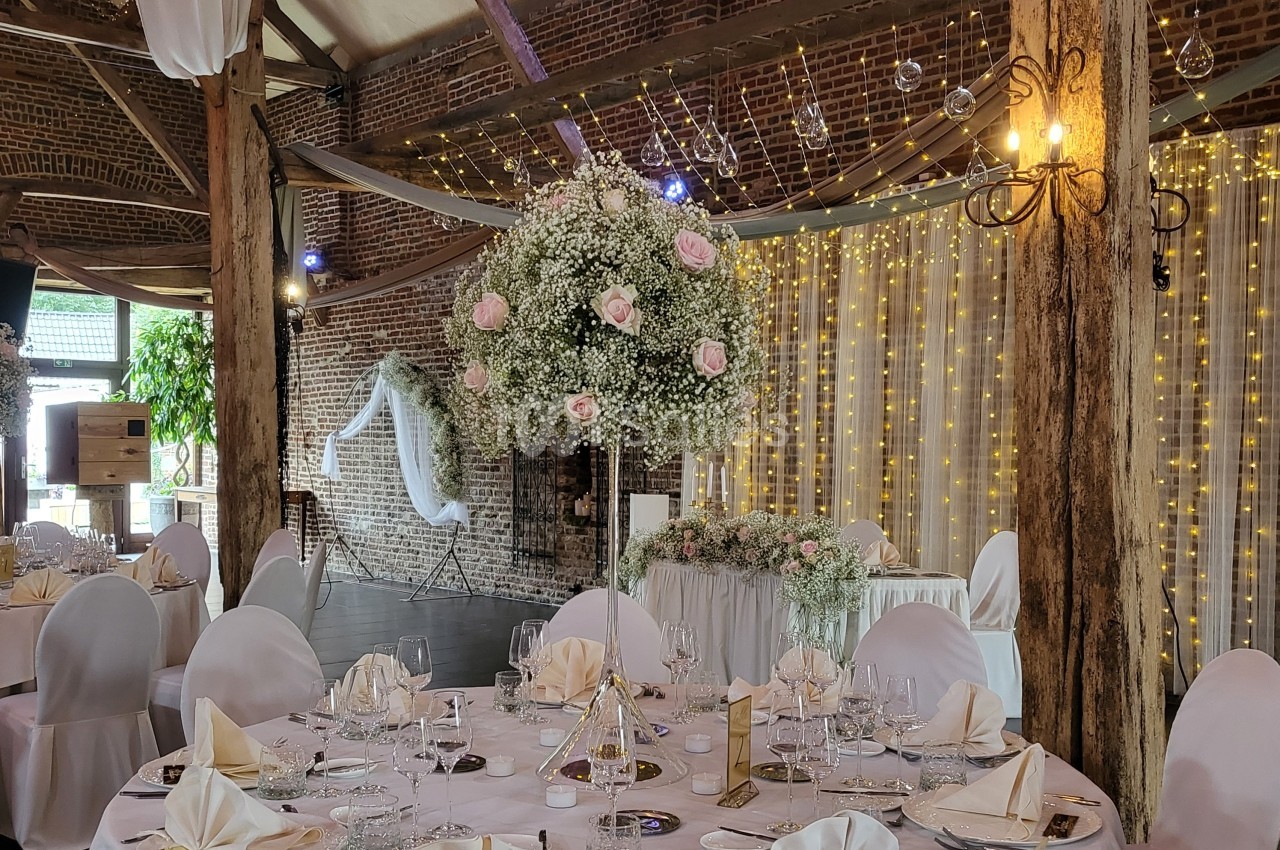 Salle de réception décorée avec des tables dressées, un centre de table floral et des guirlandes lumineuses en arrière-plan.