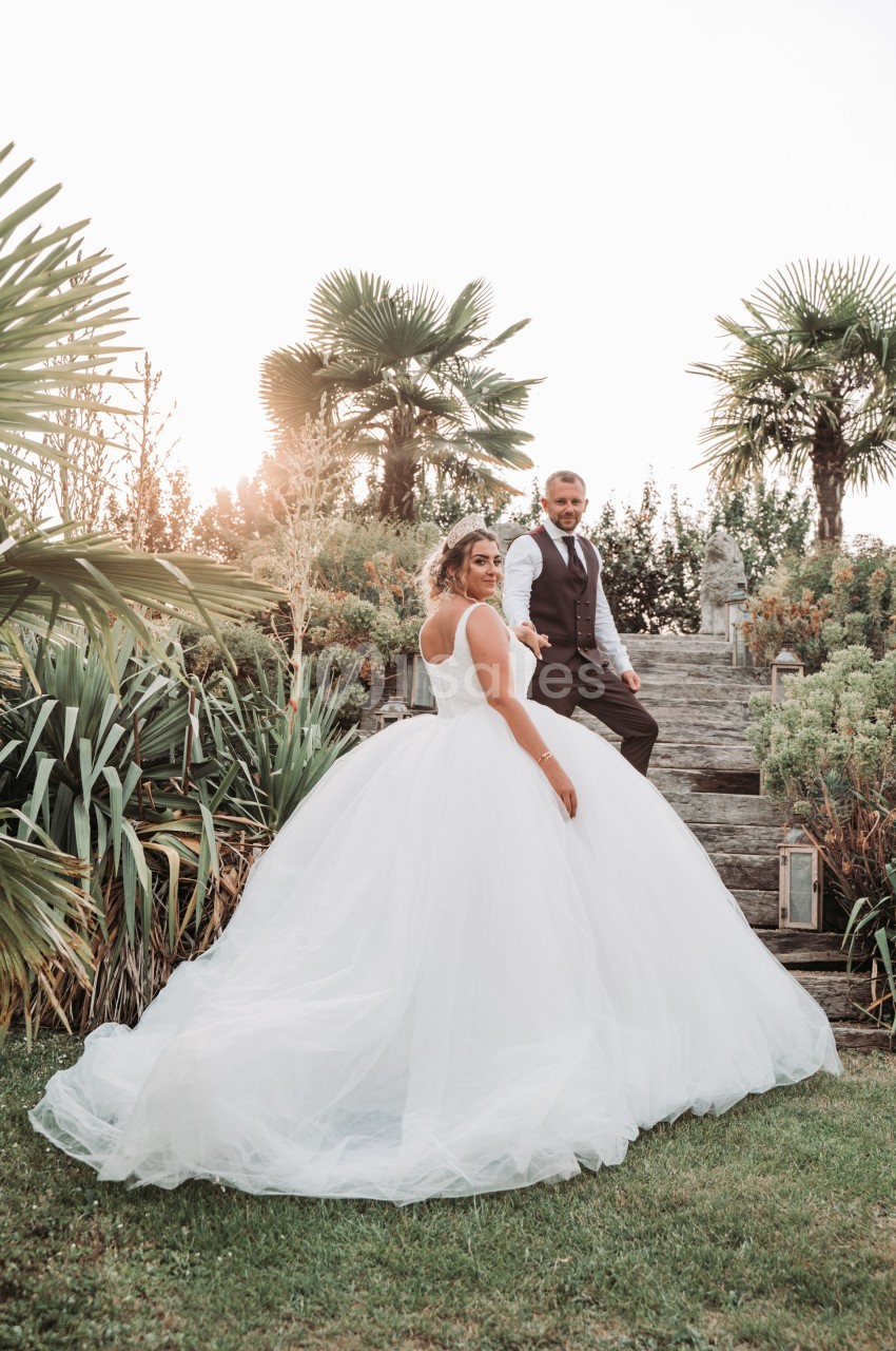 Un couple en tenue de mariage pose dans un jardin verdoyant avec des palmiers et des escaliers en pierre.