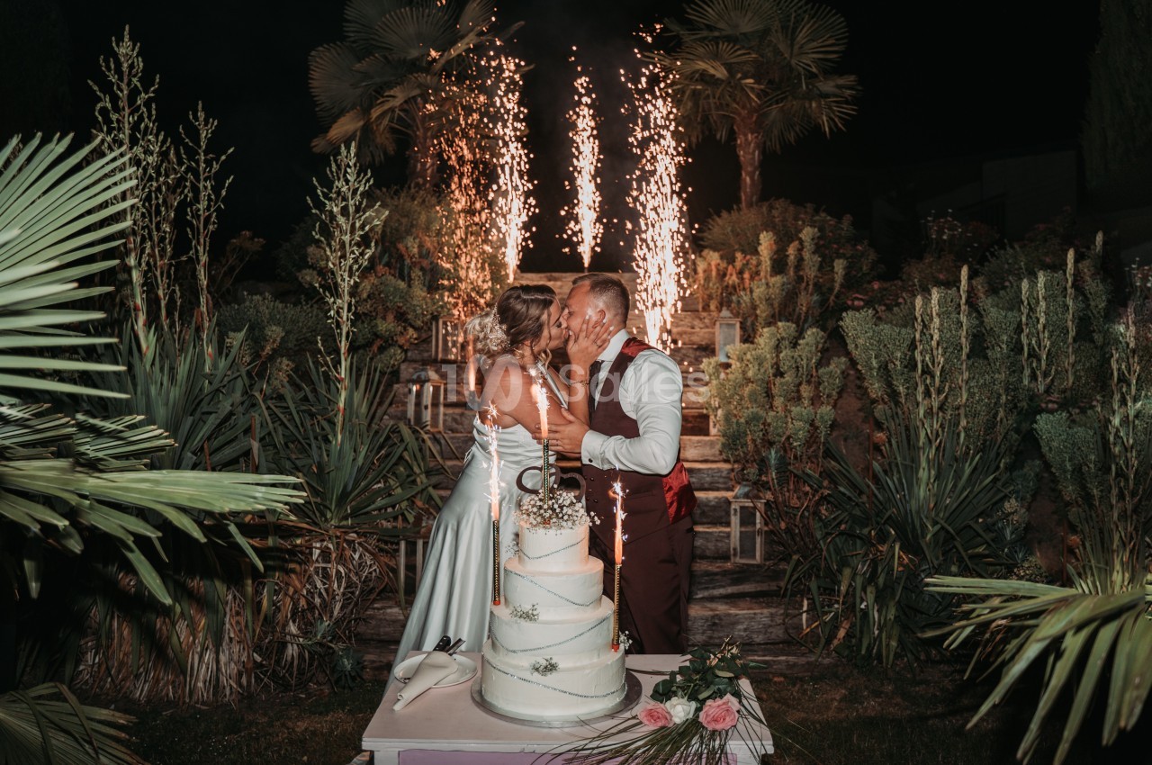 Un couple s'embrasse devant un gâteau de mariage, entouré de végétation et de feux d'artifice en arrière-plan.