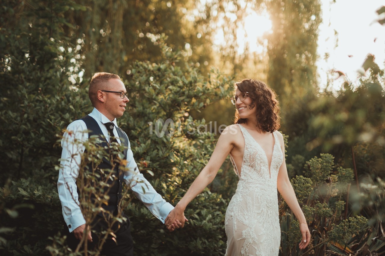 Un couple en tenue de mariage se tient la main dans un jardin baigné par une lumière douce au coucher du soleil.