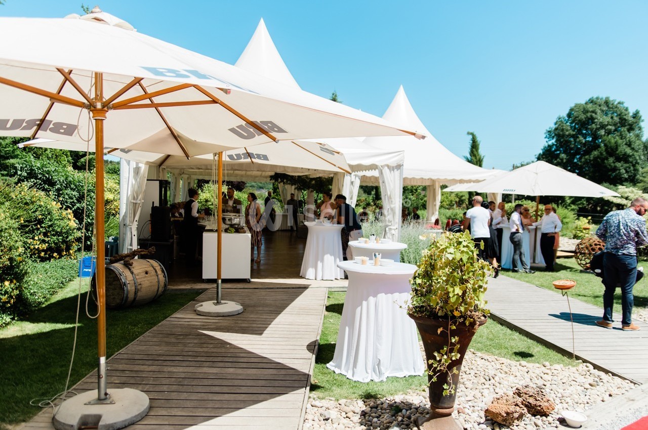 Espace extérieur avec tentes blanches, parasols, tables hautes et invités lors d'un événement en plein air.