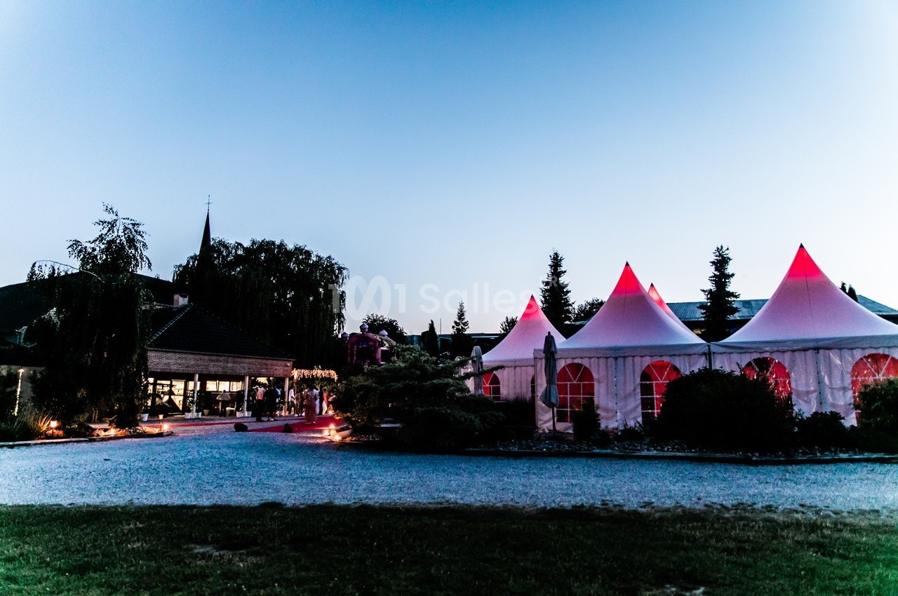 Tentes blanches éclairées en rouge dans un jardin au crépuscule, avec un bâtiment et des arbres en arrière-plan.