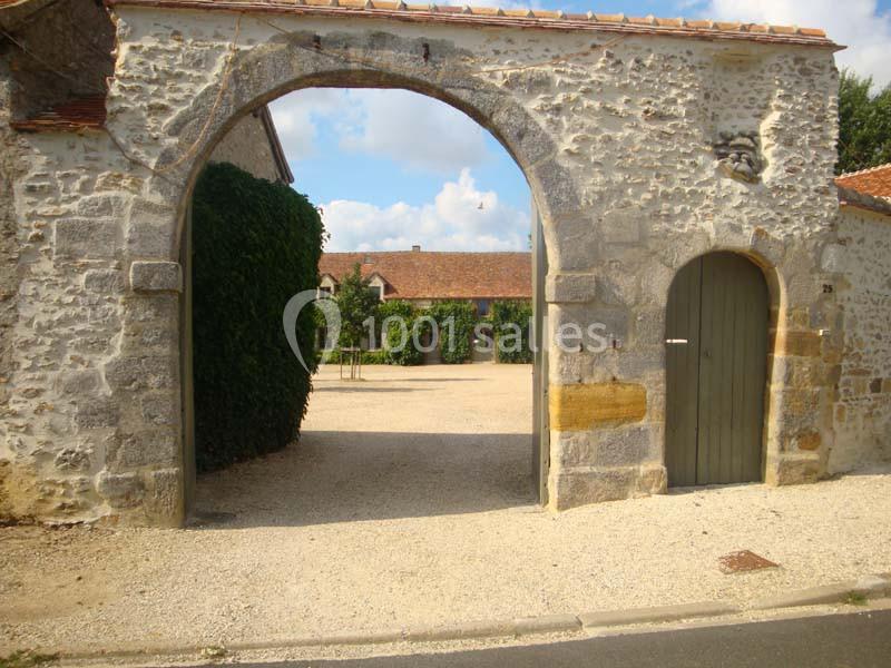 Porte en pierre voûtée donnant sur une cour gravillonnée avec des bâtiments en pierre et toits en tuiles.