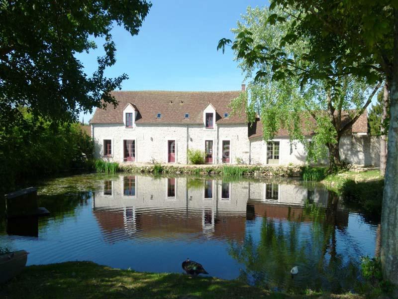 Maison en pierre avec toit en tuiles, entourée d'arbres, reflétée dans un étang calme par une journée ensoleillée.