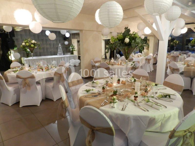 Salle de réception décorée avec des tables rondes, nappes blanches, centres de table fleuris et lanternes suspendues.