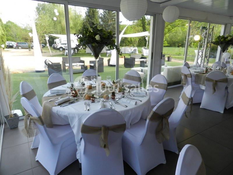 Salle de réception lumineuse avec tables rondes dressées, nappes blanches et chaises décorées de rubans beiges.
