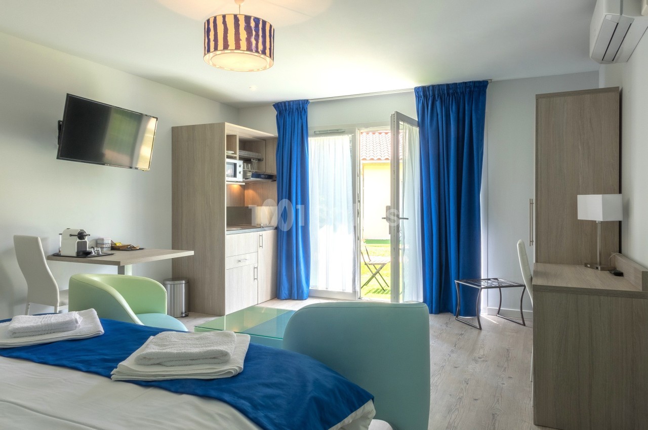Chambre lumineuse avec lit double, kitchenette, bureau, télévision murale et porte-fenêtre donnant sur un espace extérieur.