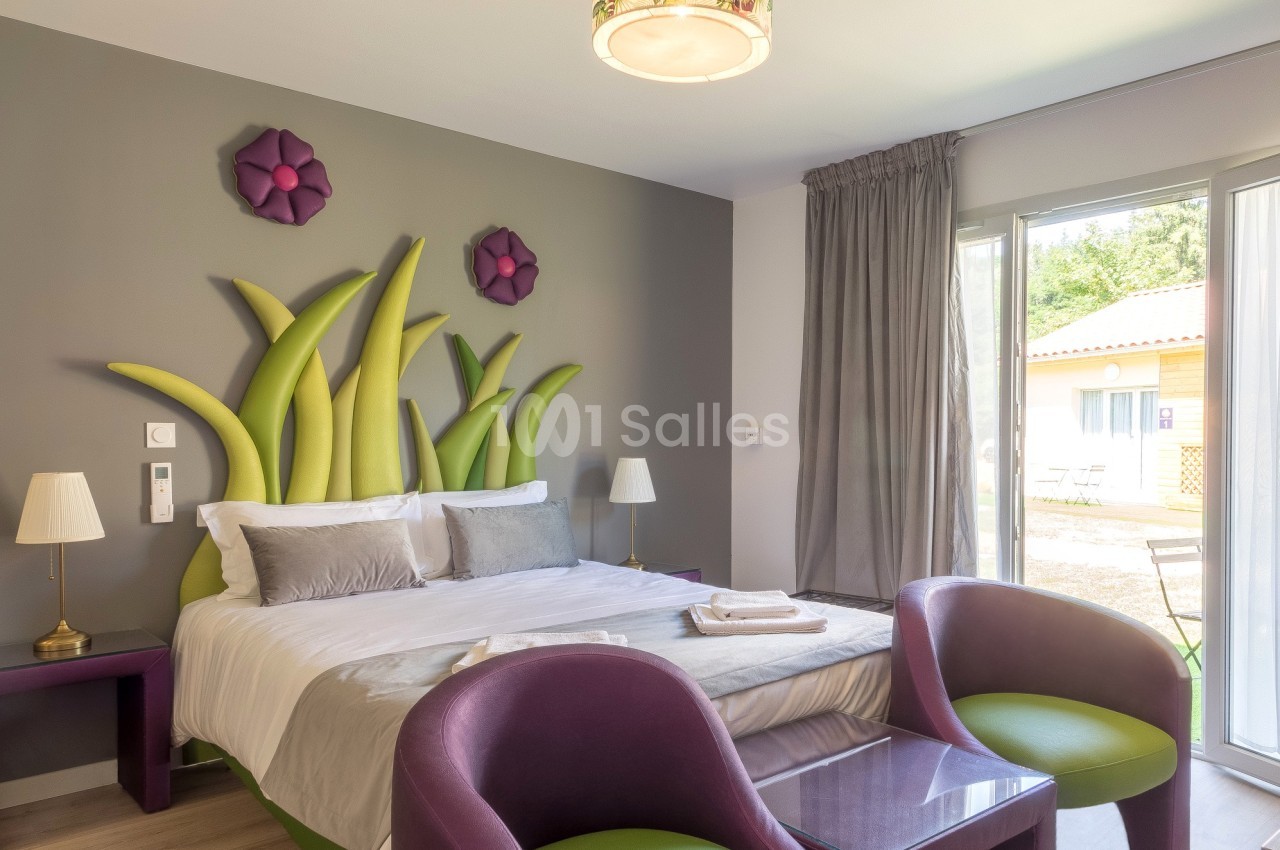 Chambre lumineuse avec lit double, décoration murale végétale, fauteuils violets et vue sur l'extérieur par une baie vitrée.