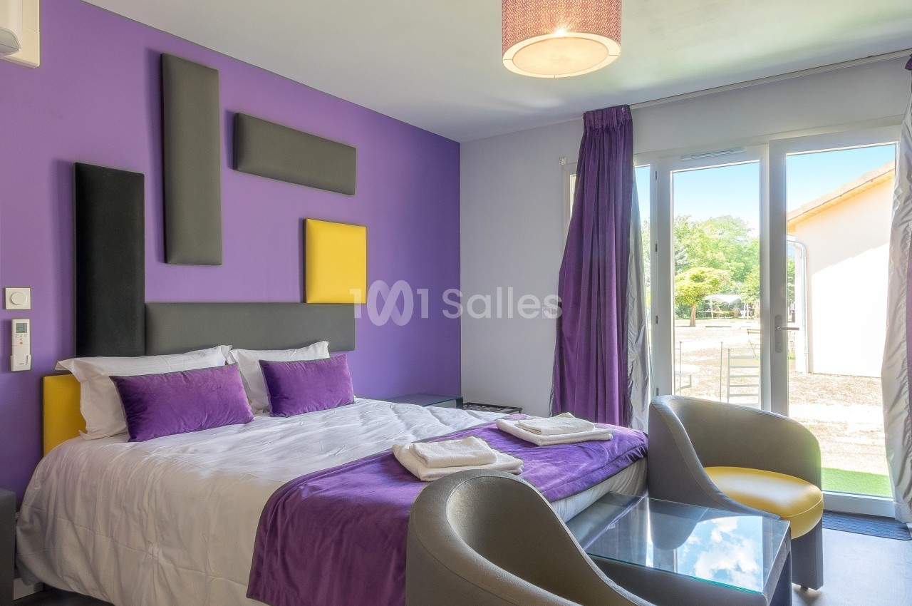 Chambre lumineuse avec lit double, murs violets, fauteuils gris, table basse en verre et grande baie vitrée donnant sur l…
