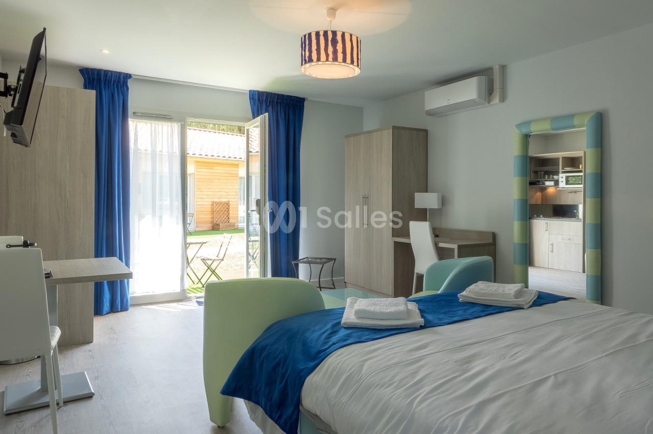 Chambre lumineuse avec lit double, bureau, kitchenette et accès à une terrasse extérieure par une porte vitrée.