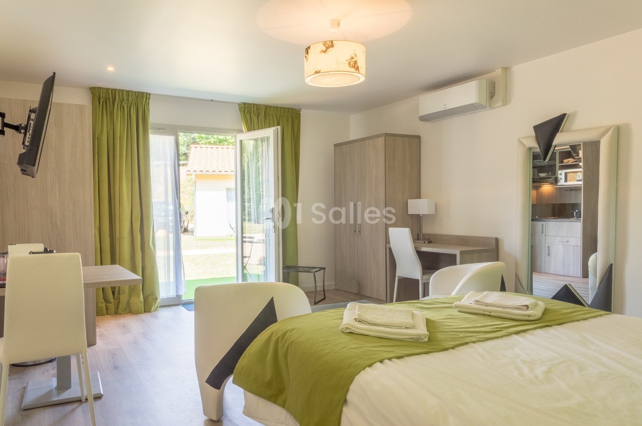Chambre lumineuse avec lit double, bureau, miroir, télévision, et porte-fenêtre donnant sur un espace extérieur.