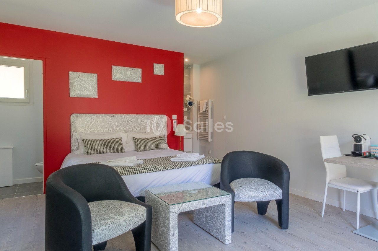 Chambre moderne avec un mur rouge, lit double, coin salon, bureau, télévision murale et salle de bain attenante.