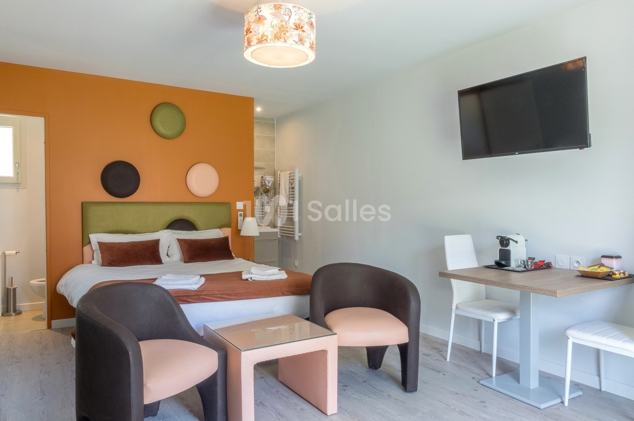 Chambre moderne avec lit double, coin salon, bureau, télévision murale, et salle de bain attenante visible en arrière-plan.