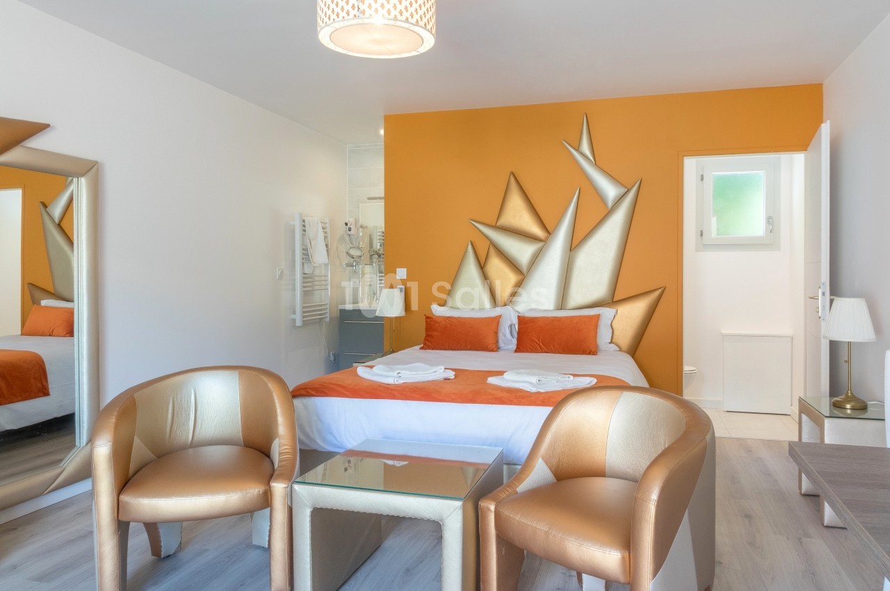 Chambre moderne avec lit double, mur orange décoratif, deux fauteuils dorés et lumière naturelle.