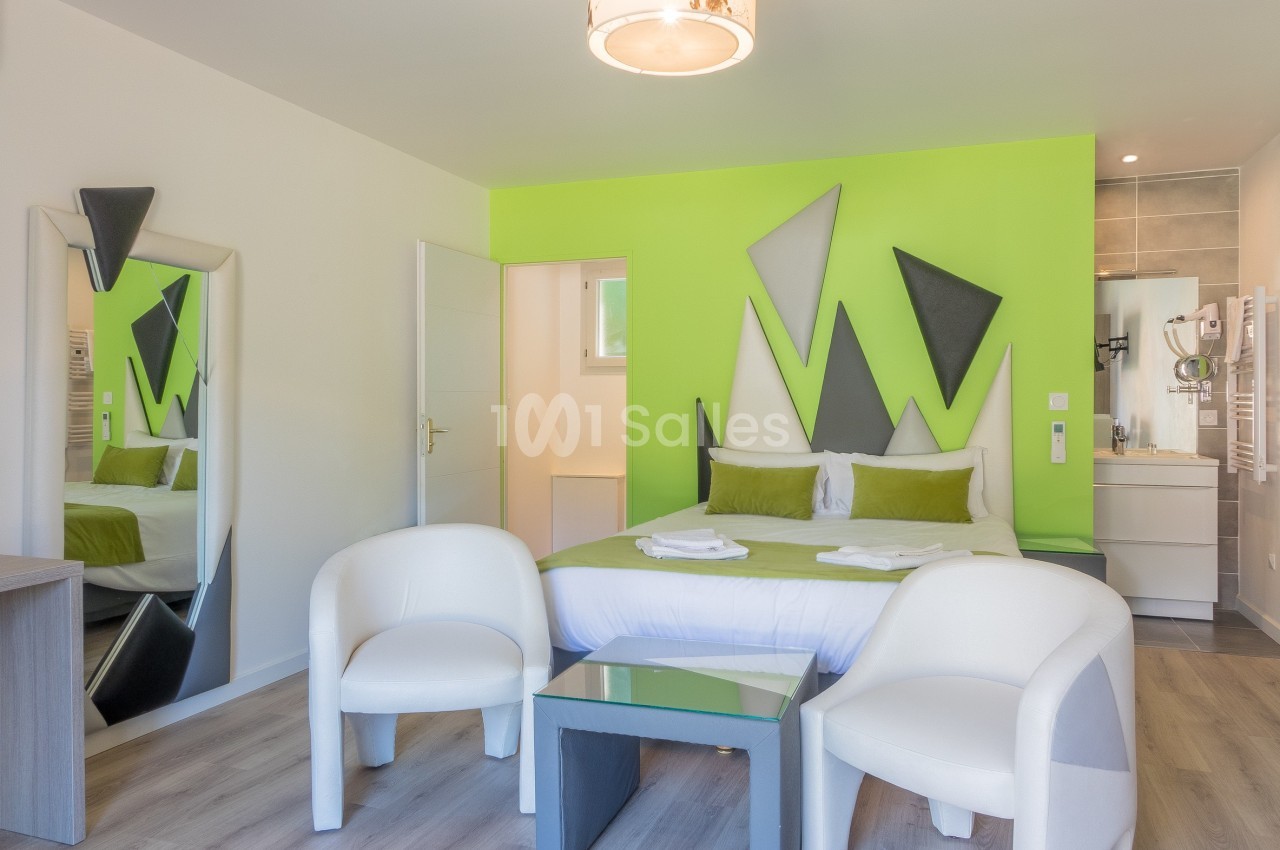 Chambre moderne avec lit double, mur vert décoré, deux fauteuils blancs, miroir et salle de bain attenante.